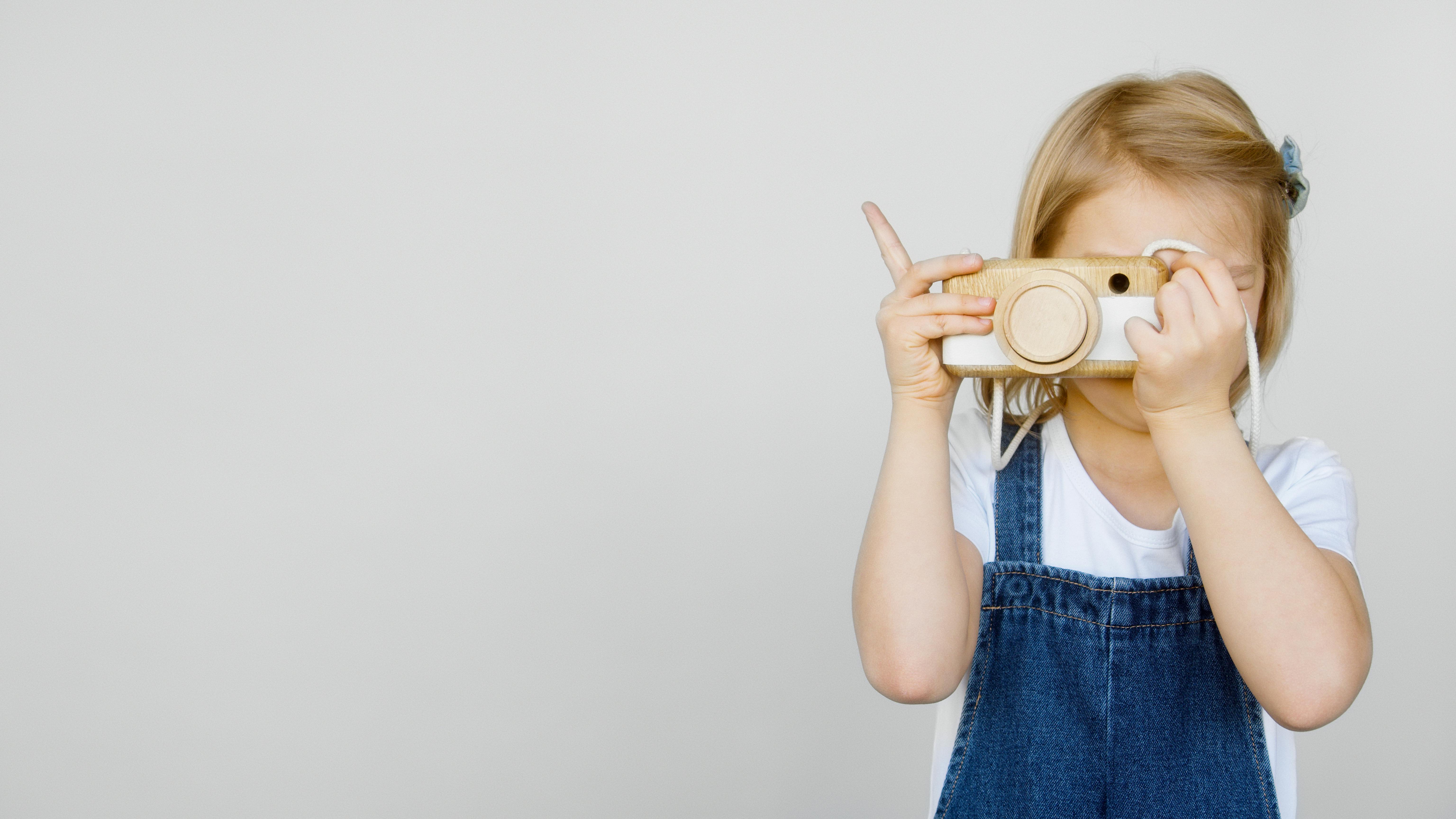 A young girl in denim overalls holding a toy camera against a plain white background.